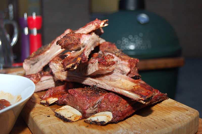 Im Vordergrund sind gegrillte Spareribs zu sehen, im Hintergrund einer der besten Keramikgrills: Der Big Green Egg XLARGE.